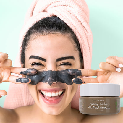 Young woman smiling with dead sea mud mask applied to her face and a jar of Deluvia Hydrating Dead Sea Mud Mask with Aloe in the foreground