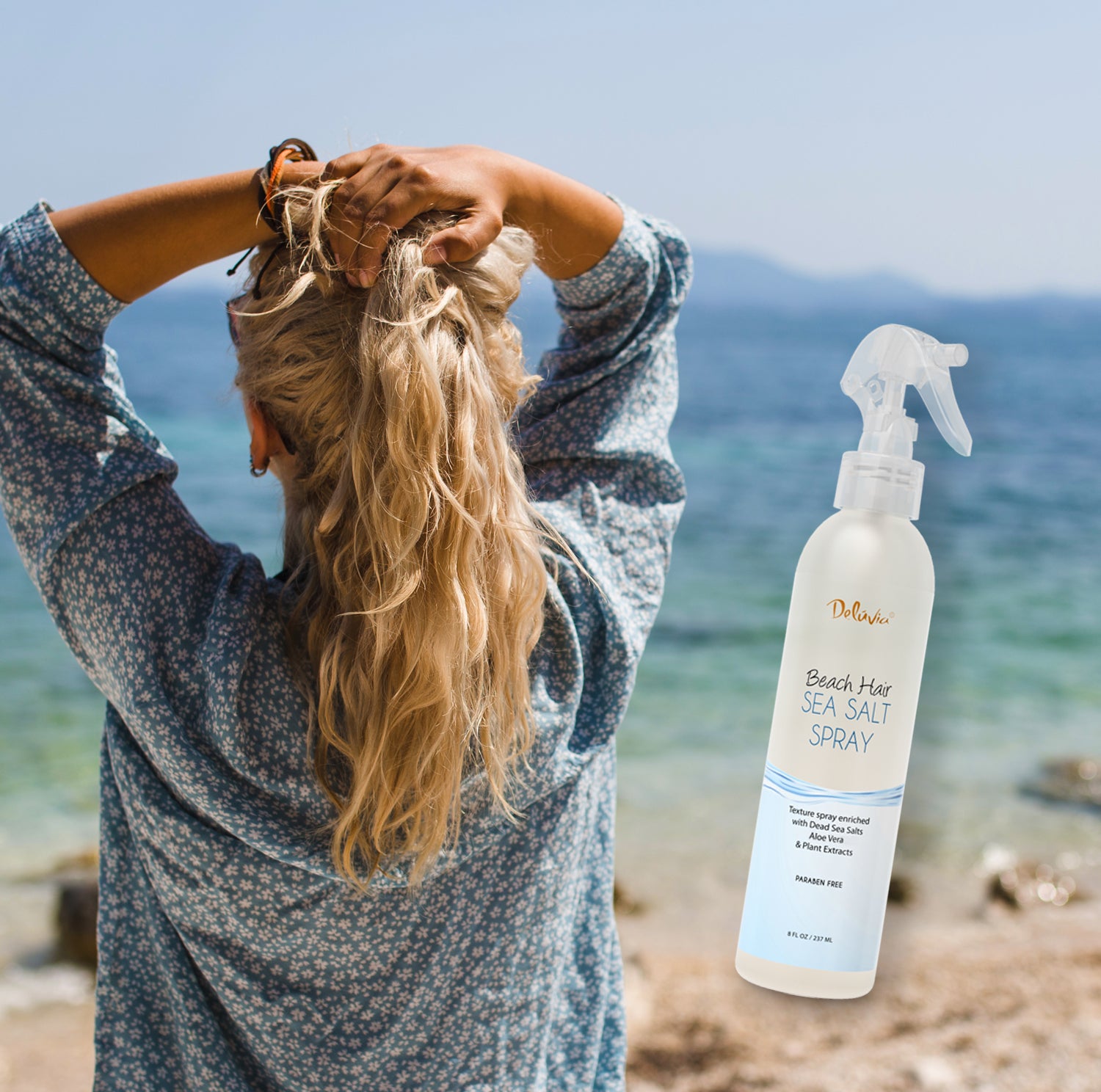 Photocomposition showing a young blonde woman facing the sea and a rocky beach in daylight, with an inclusion of a spray bottle of Deluvia's Beach Hair Sea Salt Spray