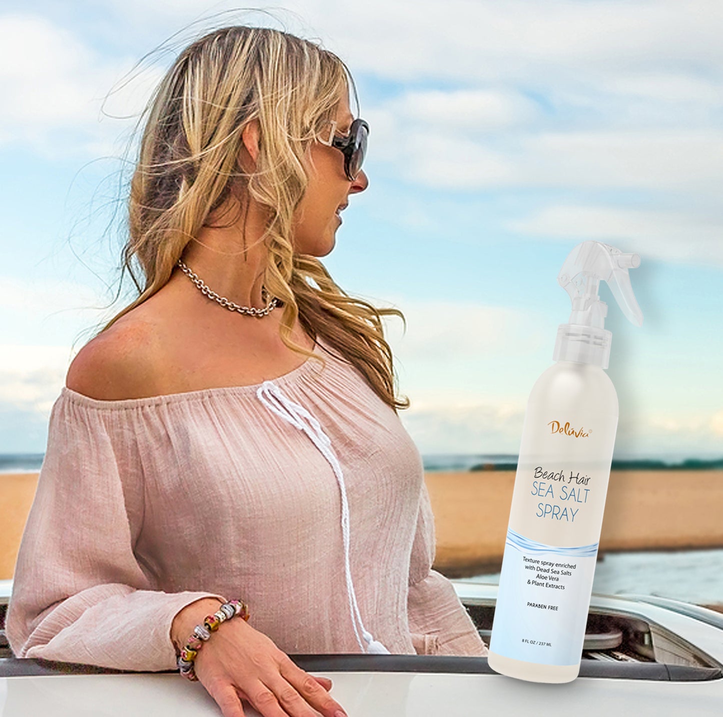 Photocomposition showing a young blonde woman with sunglasses facing a sand bar and the sea during daylight, with an inclusion of a spray bottle of Deluvia's Beach Hair Sea Salt Spray